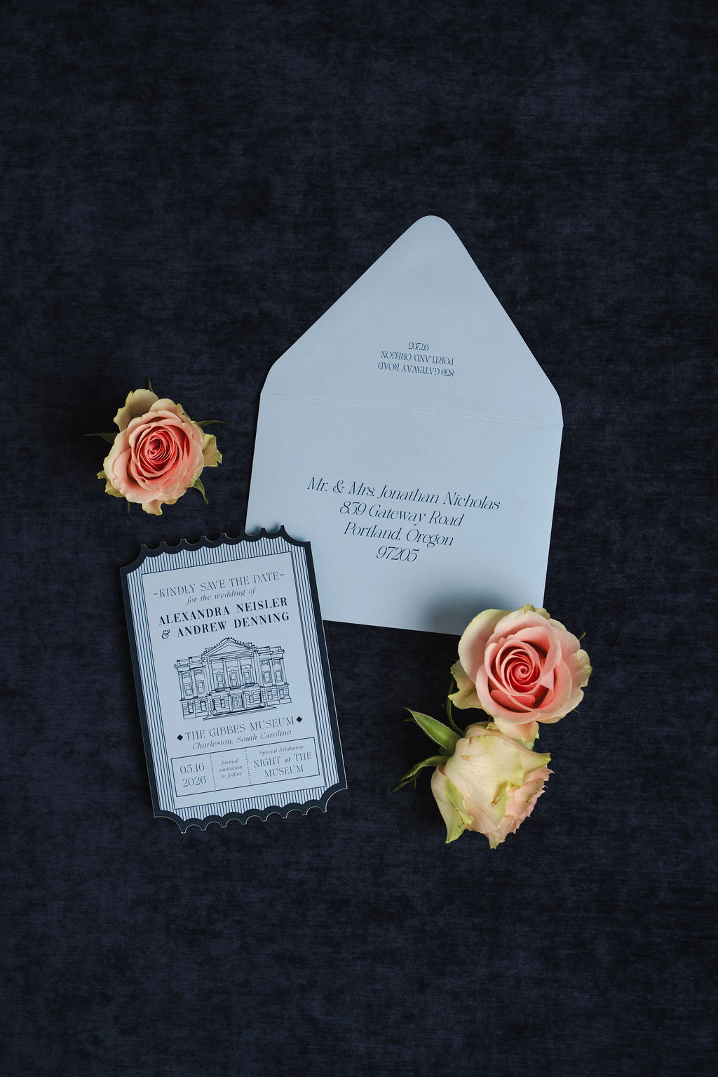 Wedding save-the-date card with a matching envelope featuring elegant navy typography, arranged with peach roses on a dark navy fabric background.